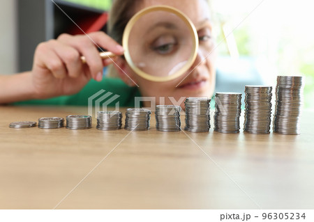 Woman looks through magnifying glass at different stacks of coins on table. Woman looks through magnifying glass at different stacks of coins on table. 96305234