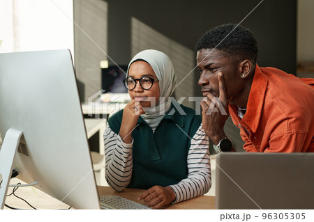 Two young serious intercultural IT engineers looking at data on computer screen or waiting for web page uploading at meeting in office 96305305
