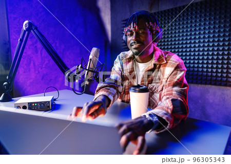 African radio host sitting at desk recording in studio with microphone and laptop 96305343