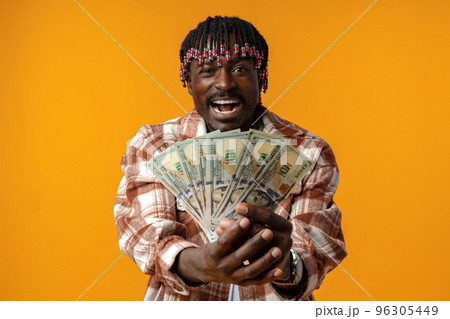 Young happy rich african man in casual shirt holding dollar bills against yellow background Young happy rich african man in casual shirt holding dollar bills against yellow background 96305449