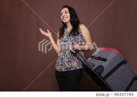 Smiling indian traveler standing with rolling suitcase, chatting, going on vacation. Woman holding baggage on wheels, cheerful tourist talking, carrying luggage in airport, trip concept 96307784