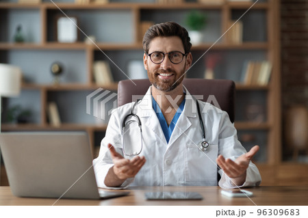 Medical Consultation. Handsome Male Doctor Sitting At Desk And Talking At Camera Medical Consultation. Handsome Male Doctor Sitting At Desk And Talking At Camera 96309683
