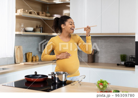 Smiling young black pregnant lady with big belly prepares dinner, tastes soup in minimalist kitchen interior 96309780