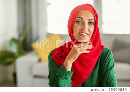 Happy middle aged muslim woman wearing red hijab, posing and smiling at camera, sitting at home in living room interior Happy middle aged muslim woman wearing red hijab, posing and smiling at camera, sitting at home in living room interior 96309880