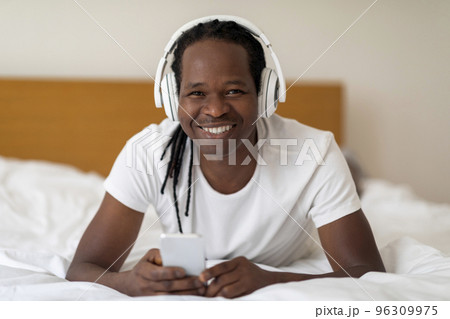 Happy Young Black Man Wearing Wireless Headphones Relaxing In Bed With Smartphone 96309975