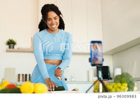 Excited black fit woman in sportswear recording video on smartphone camera, preparing vegetables salad in kitchen Excited black fit woman in sportswear recording video on smartphone camera, preparing vegetables salad in kitchen 96309983