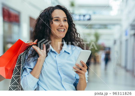 Attractive and satisfied young Hispanic woman shopping in a shopping mall, holding colorful bags and a mobile phone. Calls, types messages, pays for online purchases. 96311826