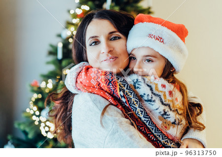 Portraits of smiling mother hugging her smiling little daughter in santa hat on Christmas tree background. Family holidays. Festive christmas mood. Happy childhood. Selective focus. Portraits of smiling mother hugging her smiling little daughter in santa hat on Christmas tree background. Family holidays. Festive christmas mood. Happy childhood. Selective focus. 96313750