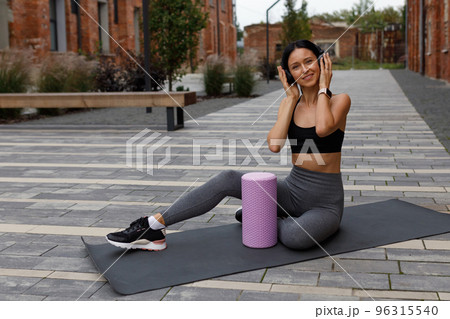 Portrait of cheerful fitness woman in headphones listening music while sitting on yoga mat outdoors 96315540