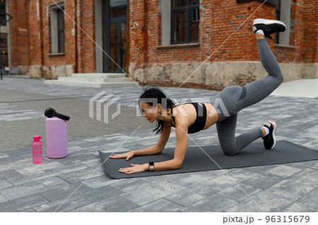 Fitness woman in sportswear doing donkey kick exercise with leg raise on yoga mat outdoor 96315679