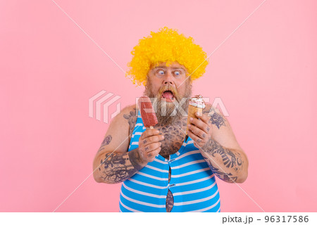 Fat man with beard and wig eats a popsicle and an icecream Fat man with beard and wig eats a popsicle and an icecream 96317586