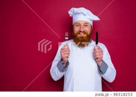 happy chef with beard and red apron holds cutlery in hand 96318253