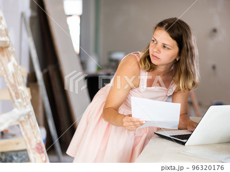Young attractive girl checks the quality and correctness of the completed repairs using a laptop inside cottage Young attractive girl checks the quality and correctness of the completed repairs using a laptop inside cottage 96320176