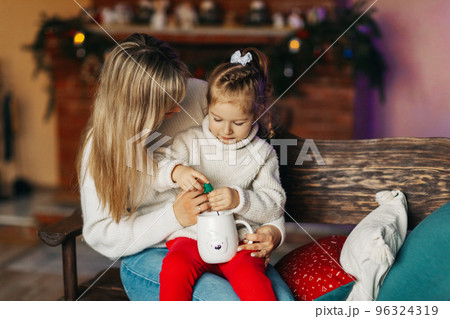 Portrait of a happy little girl sitting on her mother's lap and drinking hot chocolate on Christmas or New Year's Eve. Christmas mood, Christmas spirit, time together, motherhood, childhood 96324319