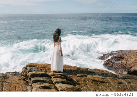 Aerial Drone View of a Bride, Brunette Girl in White Wedding Dress on Cliff Against Sea Background Aerial Drone View of a Bride, Brunette Girl in White Wedding Dress on Cliff Against Sea Background 96324321