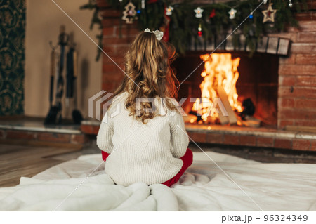 A happy little girl is sitting by the fireplace in a cozy living room on Christmas Eve. The concept of Christmas and New Year. Rear view A happy little girl is sitting by the fireplace in a cozy living room on Christmas Eve. The concept of Christmas and New Year. Rear view 96324349