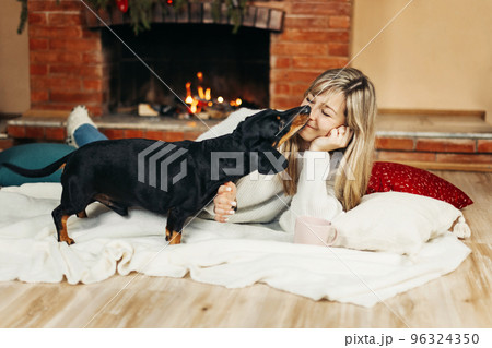A smiling young woman is playing with a dog, sitting on the floor near the fireplace, on Christmas Eve. Christmas mood, Christmas holidays. The concept of Christmas and New Year A smiling young woman is playing with a dog, sitting on the floor near the fireplace, on Christmas Eve. Christmas mood, Christmas holidays. The concept of Christmas and New Year 96324350