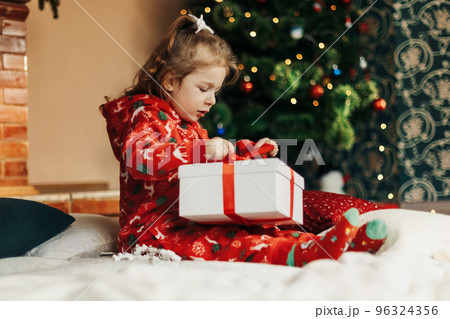 A little girl opens a Christmas present near the Christmas tree. Christmas time, gifts, festive mood. The concept of Christmas and New Year A little girl opens a Christmas present near the Christmas tree. Christmas time, gifts, festive mood. The concept of Christmas and New Year 96324356