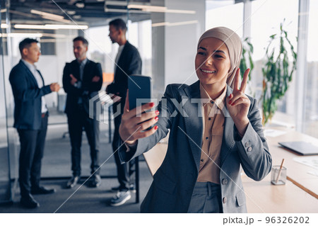 Smiling muslim business woman in hijab taking a selfie on colleagues background in office Smiling muslim business woman in hijab taking a selfie on colleagues background in office 96326202