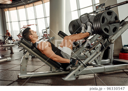 Man Using A Press Machine In A Fitness Club. Strong man doing an exercise on its feet in the simulator. Side view. Man Using A Press Machine In A Fitness Club. Strong man doing an exercise on its feet in the simulator. Side view. 96330547