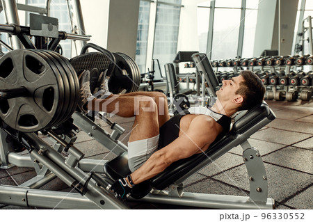 Man Using A Press Machine In A Fitness Club. Strong man doing an exercise on its feet in the simulator. Side view. Man Using A Press Machine In A Fitness Club. Strong man doing an exercise on its feet in the simulator. Side view. 96330552