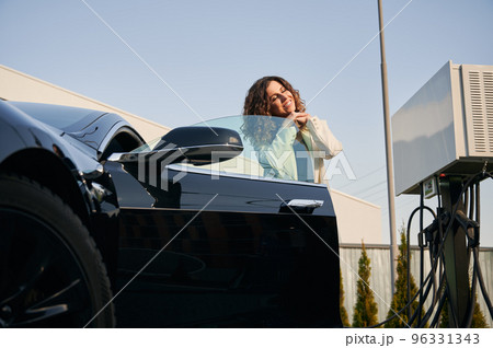 Beautiful businesswoman in stylish suit standing by electric car on the street at vehicle charging station. Joyful woman opening automobile door and smiling while posing at electric charging station. 96331343