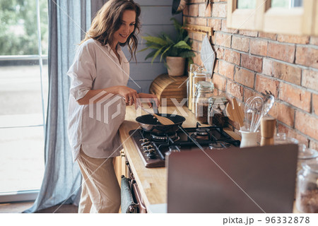 Woman stirring food cooking in a frying pan on the stove and looking at her laptop, she reading cooking recipe or watching show while frying food. Woman stirring food cooking in a frying pan on the stove and looking at her laptop, she reading cooking recipe or watching show while frying food. 96332878