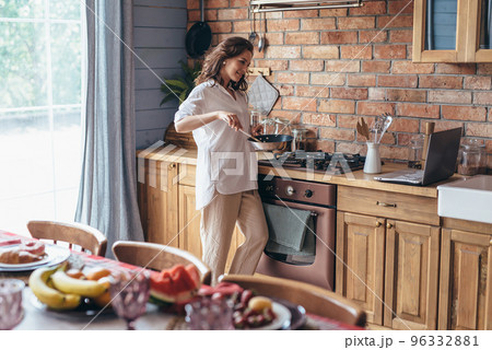 Woman in the kitchen cooking in a frying pan and looking at her laptop screen. Woman in the kitchen cooking in a frying pan and looking at her laptop screen. 96332881