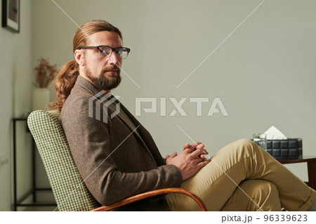 Portrait of mature bearded professional psychotherapist in eyeglasses looking at camera sitting on armchair during his work at office 96339623