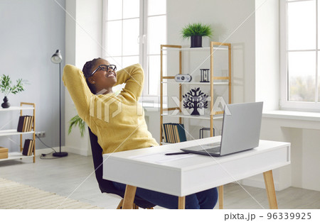 Smiling young woman sitting in front of laptop with arms behind head 96339925