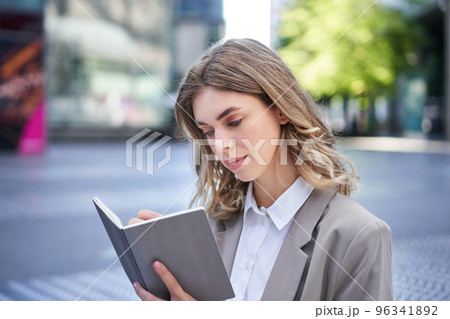 Businesswoman in corporate suit, writes down smth, works on ideas in notebook, sits outside in city centre Businesswoman in corporate suit, writes down smth, works on ideas in notebook, sits outside in city centre 96341892