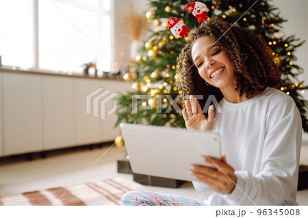 A young woman on the background of a Christmas tree with gifts, with a tablet has a video call or video chat. 96345008