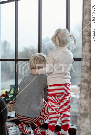Back view on brother and sister on Christmas Eve near window. Little boy and girl in red striped pants peek out window. 96345076