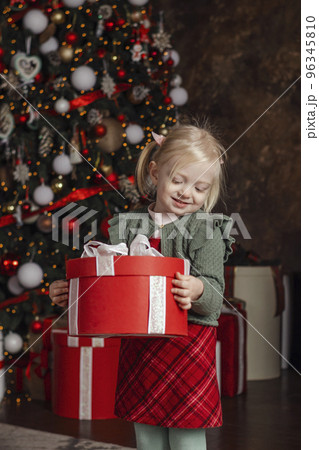 Little happy smiling girl with Christmas gift box on xmas tree background. Traditional xmas present. Vertical frame Little happy smiling girl with Christmas gift box on xmas tree background. Traditional xmas present. Vertical frame 96345810