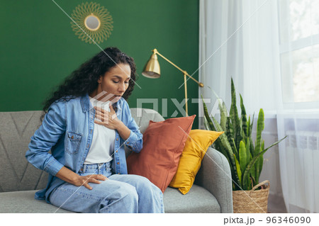 Panic attack. A young beautiful Latin American woman is sitting at home on the sofa, holding her chest with her hand, breathing heavily. Mental health problems, needs help. Panic attack. A young beautiful Latin American woman is sitting at home on the sofa, holding her chest with her hand, breathing heavily. Mental health problems, needs help. 96346090