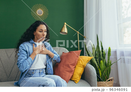 A young Latin American asthmatic woman is experiencing an asthma attack. He is sitting at home on the sofa, holding his chest with his hands, it is difficult to breathe, he needs medical help. 96346091