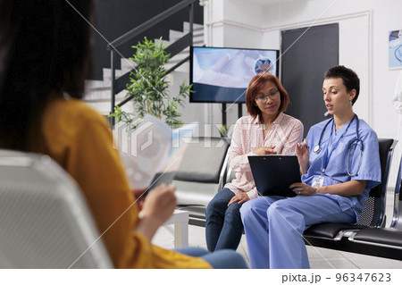 Health specialist consulting old patient in hospital lobby, taking notes on digital device and filling in report papers. Nurse doing checkup examination with senior woman in waiting room. 96347623