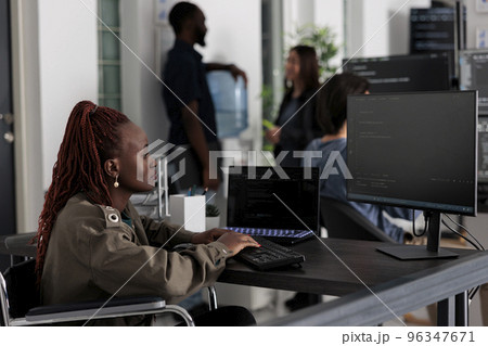 Programmer in wheelchiar typing html code on computer, working with cloud computing terminal window. Disabled software developer writing artificial intelligence algorithm in it company office. 96347671
