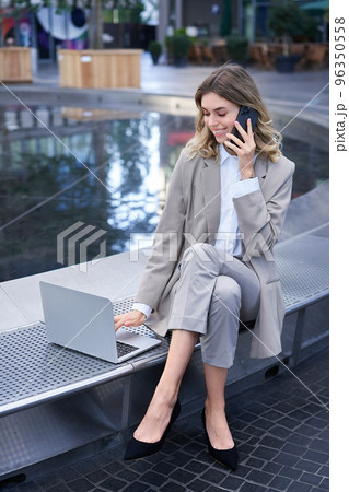 Vertical shot of corporate woman sitting outdoors with laptop, talking on mobile phone, working ouside office building while waiting for someone 96350558