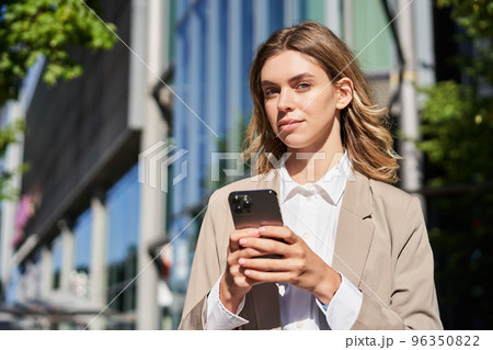 Portrait of stylish businesswoman in corporate outfit, holding smartphone, standing on street and using mobile phone in hands Portrait of stylish businesswoman in corporate outfit, holding smartphone, standing on street and using mobile phone in hands 96350822