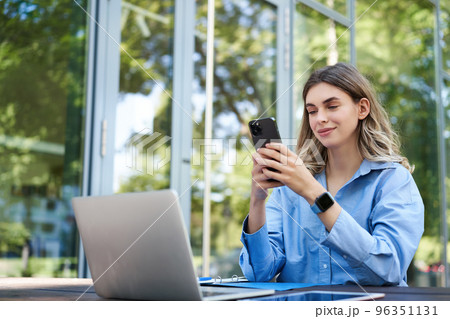 Portrait of saleswoman working outside office, sititng with laptop, using mobile phone. Digital nomad woman studying, using computer outdoors 96351131