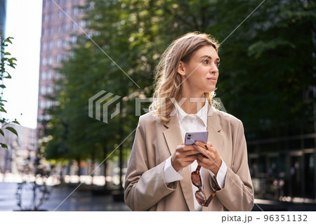 Portrait of businesswoman in suit, holds her mobile phone while walking on street 96351132