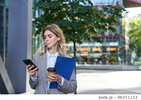 Businesswoman drinking coffee and holding documents with smartphone, going to work in office, standing outdoors in city center 96351133