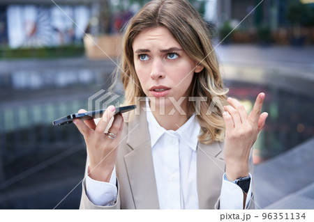 Portrait of confused businesswoman recording voice message, counting, thinking while talking into mobile phone, sitting near ofice buildings on street 96351134