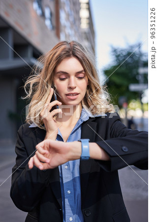 Portrait of confident businesswoman going on a meeting, standing on street, talking on mobile phone and checking time on digital watch Portrait of confident businesswoman going on a meeting, standing on street, talking on mobile phone and checking time on digital watch 96351226