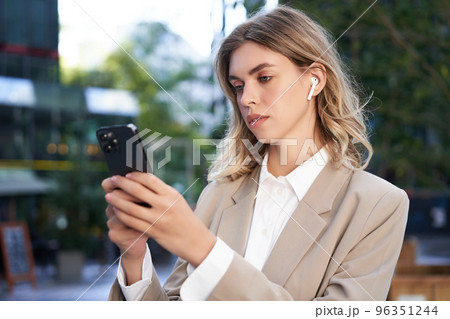 Successful businesswoman in beige suit, wireless headphones, looking at mobile phone, using smartphone app, standing outdoors on street Successful businesswoman in beige suit, wireless headphones, looking at mobile phone, using smartphone app, standing outdoors on street 96351244