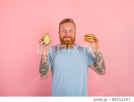 Happy man with beard and tattoos is undecided if to eat an avocado or an hamburger 96352247