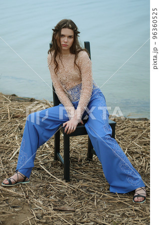 Full length portrait of young woman in blue pants and lace blouse sitting on chair shore of the lake 96360525