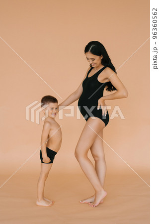 Happy motherhood. A pregnant woman in the studio on a beige background with her five-year-old son, measuring the size of their bellies Happy motherhood. A pregnant woman in the studio on a beige background with her five-year-old son, measuring the size of their bellies 96360562