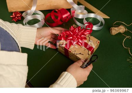 Overhead view of woman's hands tying beautiful bow from a shiny red ribbon while decorating a Christmas gift. Wrapping. Overhead view of woman's hands tying beautiful bow from a shiny red ribbon while decorating a Christmas gift. Wrapping. 96369967